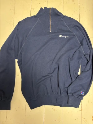 Vintage Blå half zip sweatshirt från Champion - Snygg vintage marinblå sweatshirt från Champion med half zip och hög krage. Tröjan har broderad logga på bröstet och ett litet Champion-märke på ärmen. Tillverkad i mjuk bomull, perfekt för chill dagar. Långärmad och med ribbade muddar för extra komfort. Köpt second hand. Sjukt nice och den sitter baggy. 