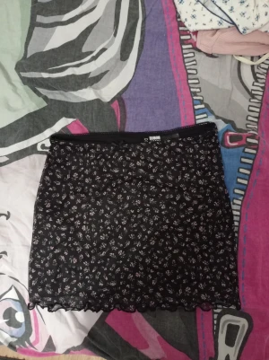 Svart blommig kortkjol från H&M Divided - Supersöt svart kortkjol från H&M Divided med små blommor i vitt och ljusrosa. Kjolen har en tight passform, volangkant nertill och är gjord i mjuk polyester med lite stretch. Perfekt för dig som gillar en feminin och trendig look.