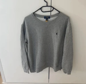 Grå sweatshirt från Polo Ralph Lauren - Snygg grå sweatshirt från Polo Ralph Lauren med klassisk rund hals och broderad marinblå logga på bröstet. Tröjan har ribbade muddar vid ärmslut och nederkant, perfekt för chill dagar. Mjuk insida i bomullsmix som känns skön mot huden.