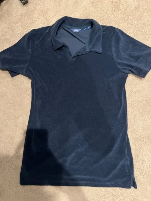 Mörkblå frottéskjorta Jack & Jones - Mörkblå skjorta från Jack & Jones i mjuk frotté. Klassisk krage och korta ärmar ger en chill och avslappnad vibe. Perfekt för dig som gillar stilren design utan massa mönster. Skön att bära och enkel att matcha med jeans eller shorts. Storlek 176.