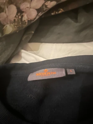 Svart sweatshirt från Morris, storlek L - Svart sweatshirt från Morris i storlek L. Tröjan har rund hals och lång ärm, med en liten guldfärgad logga broderad på bröstet. Perfekt för dig som gillar stilrena och klassiska plagg med diskreta detaljer.