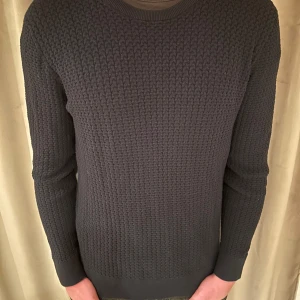 J.lindberg tröja - Säljer en stilren mörkgrå stickad tröja med rund hals och långärmad design. Perfekt för kyligare dagar med sitt klassiska mönster och bekväma passform.