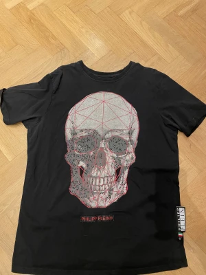 Svart t-shirt med döskalle från Philipp Plein - Svart t-shirt från Philipp Plein med stor döskalle framtill i silver och röda linjer, dekorerad med små svarta pärlor. Kortärmad modell i mjukt material, med logotyp på bröstet och sidan. Riktigt edgy och unik design. Tröjan har tecken på användning då paljetter har lossnat.