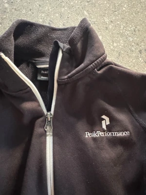 Svart fleecejacka Peak Performance - Svart fleecejacka från Peak Performance med vit dragkedja och broderad logga på bröstet. Jackan har hög krage, långa ärmar och två fickor med dragkedja. Perfekt för kyliga dagar och enkel att slänga på sig när du är på språng.