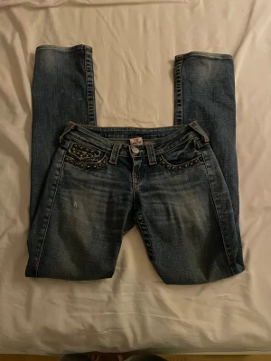 True Religion bootcut jeans med nitar - Snygga blå jeans från True Religion i bootcut-modell med coola nitar och detaljer på fickorna både fram och bak. Jeansen har klassisk femficksdesign, ljus tvätt och kontrastsömmar. Perfekt för dig som vill sticka ut med en unik look. Som tyvärr inte kommer till användning. Storleken är 26, som passar mig som har 36 i vanliga fall men även 34. 