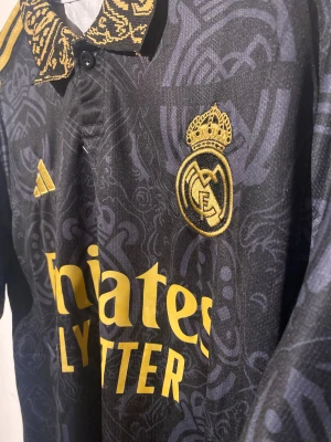 Real Madrid svart/guld matchtröja Adidas - Snygg Real Madrid fotbollströja från Adidas i svart med guldiga detaljer och klubbmärke. Tröjan har korta ärmar, mönstrad krage och ärmslut samt tryckt sponsorlogga. Tillverkad i lätt funktionsmaterial, perfekt för match eller träning. Nu under black friday veckan för endast 349 kr🔥🔥