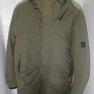 Drew parkasjacka från Didriksons - Grön parkasjacka från Didriksons med huva, stora fickor och dragkedja framtill. Jackan är vadderad. Perfekt för kalla dagar och har en clean, stilren look med diskreta detaljer.