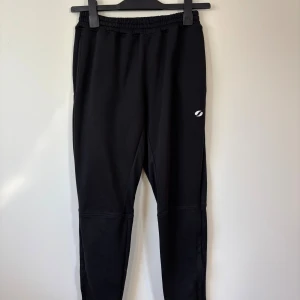 SOC träningsbyxor - 158/164 Storlek - Svarta tröningsbyxor från SOC med elastisk midja och smal passform. Byxorna har en vit logga på benet och är gjorda i mjukt syntetmaterial som är perfekt för träning. 