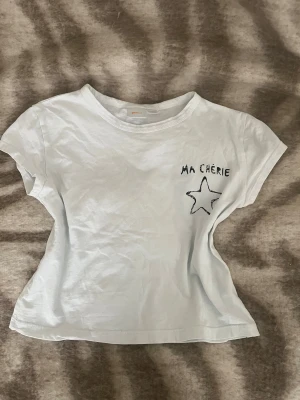 Vit t-shirt Ma Chérie med stjärna - Vit t-shirt från Gina Tricot med svart tryck 'Ma Chérie' och en handritad stjärna både fram och bak. Modellen är kortärmad och har en klassisk rund hals. T-shirten är gjord i mjuk bomull och har en avslappnad passform som är lätt att matcha. 134/140