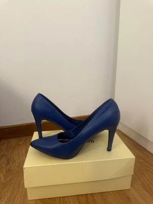 Blå pumps med spetsig tå och klack - Snygga blå pumps med spetsig tå och hög klack. Skorna har en stilren och klassisk form, tillverkade i slätt skinnliknande material. Perfekt för dig som vill sticka ut med färg och elegans. Innersulan har ett silvrigt inslag med vågig kant.