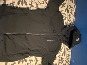 Svart pufferjacka från Sail Racing - Svart pufferjacka från Sail Racing med huva och snygga detaljer. Jackan har dragkedja framtill, flera praktiska fickor med dragkedja och logga på bröstet och huvan. Materialet är slitstark polyester och jackan är fodrad för extra värme. Perfekt för kalla dagar.