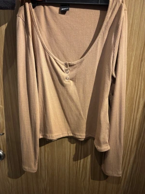 Beige ribbad långärmad topp Gina Tricot - Säljer en beige ribbad långärmad topp från Gina Tricot med rundad halsringning och tre dekorativa knappar framtill. Toppen har en croppad passform och är tillverkad i mjuk bomull som är skön mot huden. Perfekt för dig som gillar enkla och stilrena plagg.