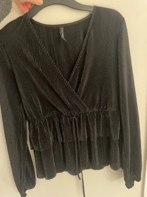 Svart blus med volanger - Säljer en svart blus med  volanger nedtill. Blusen har långa ärmar med resår  och ett knytband i midjan. Super fint skick.