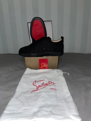 Svarta sneakers från Christian Louboutin - Säljer ett par svarta sneakers från Christian Louboutin med klassisk röd sula. Skorna har rund tå, snörning och är tillverkade i mocka med snygga svarta detaljer. Medföljer originalpåse och box. Perfekt för dig som gillar exklusiv och stilren design.