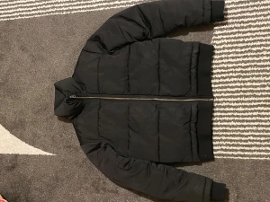 Svart pufferjacka med dragkedja - Säljer en svart pufferjacka med hel dragkedja framtill. Jackan har ribbade muddar vid ärmslut och midja, samt en hög krage. Perfekt för kalla dagar och har en clean, stilren look som passar till det mesta.