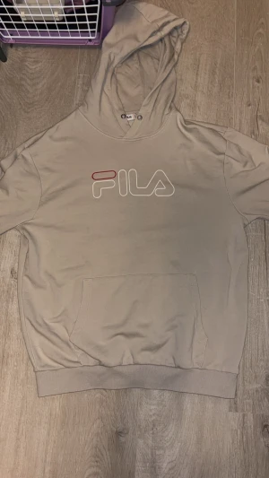 Beige hoodie från FILA med logga - Snygg beige hoodie från FILA med stor logga i vitt och rött på bröstet. Tröjan har huva med dragsko och en klassisk magficka. Perfekt för chill dagar och streetstyle. Mjuk bomullskvalitet och ribbade muddar vid ärmslut och nederkant.