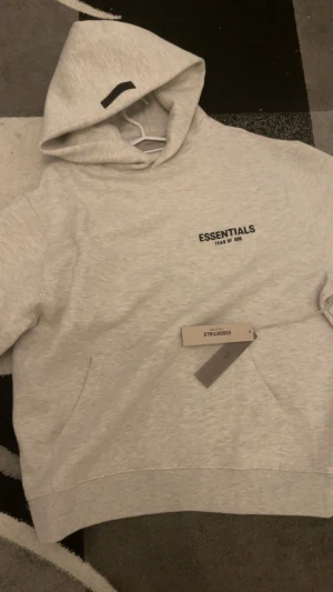 Essentials Fear of God ljusgrå hoodie - Säljer en stilren ljusgrå hoodie från Essentials Fear of God. Tröjan har huva, stor svart logga på bröstet och en klassisk känguruficka framtill. Mjuk bomullskänsla och relaxed passform, perfekt för chill dagar.