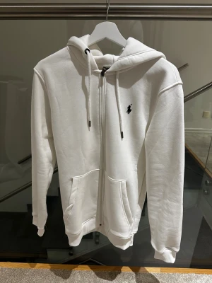 Vit kofta från Polo Ralph Lauren - Vit kofta från Polo Ralph Lauren storlek L men passar som storlek M med dragkedja och snörning i huvan. Klassisk broderad logga på bröstet i svart. Tröjan har två fickor framtill och ribbade muddar vid ärmslut och nederkant. Perfekt för chill dagar och snygg streetstyle. Skriv efter minsta lilla fundering!! Pris kan diskuteras!!