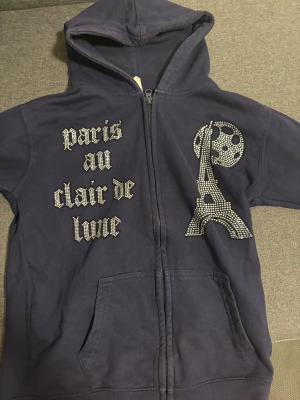 Mörkblå hoodie med strassmotiv Paris - Snygg mörkblå hoodie från BLINGLUCID med huva och dragkedja. Framsidan har strassmotiv med texten 'paris au clair de lune' och en glittrig Eiffel-torn-design. Tillverkad i mjuk bomull och polyesterblandning, perfekt för dig som gillar unika detaljer.