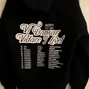 Svart Hov1 hoodie med turnétryck - Svart hoodie från Hov1 med stort vitt tryck på ryggen med texten 'Vi Dansar Vidare I Livet' och turnédatum från deras sista 2025. 