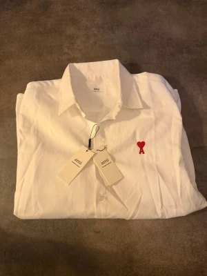 Vit skjorta från Ami med rött hjärta - Klassisk vit skjorta från Ami Alexandre Mattiussi med snyggt rött broderat hjärta och A på bröstet. Skjortan har lång ärm, knappar framtill och är gjord i mjuk bomull. Perfekt för dig som gillar stilrena plagg med en twist. XL men är som L