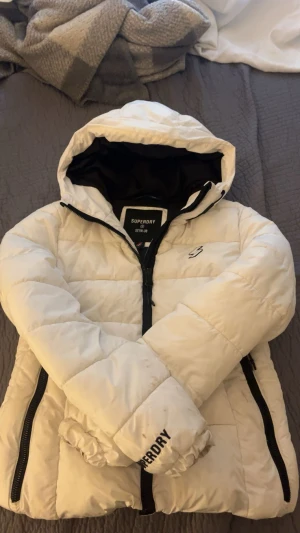 Vit pufferjacka från Superdry - Snygg vit pufferjacka från Superdry med svart dragkedja och huva. Jackan har två sidofickor med dragkedja, elastiska muddar och logga på ärmen. Perfekt för kalla dagar och ger en clean look. Passformen är normal och jackan är fodrad för extra värme.