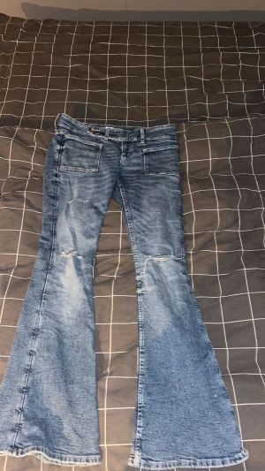 Blå bootcut jeans med hål - Säljer ett par blå jeans med bootcut passform och snygga slitningar vid knäna. Jeansen har klassiska fickor fram och bak, samt en låg midja. Perfekta för dig som gillar en retro vibe och vill sticka ut lite extra.