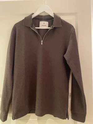 Grön långärmad tröja Jack & Jones - Säljer en brun långärmad tröja från Jack & Jones med klassisk krage och half zip-dragkedja framtill. Tröjan har en diskret struktur i tyget och är tillverkad i mjuk bomull, perfekt för dig som gillar stilren och enkel design.