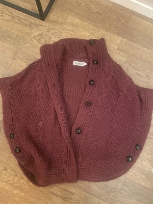 Vinröd stickad poncho från Soaked - Vinröd stickad poncho från Soaked med grovstickad struktur och dekorativa flätor. Ponchon har en hög krage och stora svarta knappar längs sidorna. Modellen är loose fit och har en rundad form som ger en chill vibe. Perfekt att slänga över en långärmad tröja för extra värme. Saknar dock en knapp.