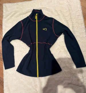 Blå fleecejacka från Kari Traa - Sportig blå fleecejacka från Kari Traa med gul dragkedja och logga på bröstet. Snygga rosa sömmar ger extra kontrast. Jackan har hög krage och långärmad passform, perfekt för träning eller chill. Materialet är mjuk fleece som håller dig varm.