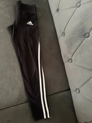 Svarta Adidas leggings med vita ränder - Snygga svarta leggings från Adidas med klassiska vita ränder längs benen och Adidas-logga på låret. Modellen är tight och har hög midja, perfekt för träning eller chill. Materialet är mjukt och stretchigt, troligtvis polyester och bomull för skön passform.