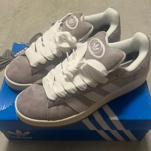 Adidas Campus grå sneakers mocka - Säljer ett par Adidas Campus sneakers i ljusgrå mocka med vita detaljer och breda vita snören. Skorna har klassiska tre ränder på sidan, Adidas-logga på plös och häl samt en gummisula i beige/brun. Perfekta för dig som gillar retrostil och streetwear. Byten funkar.