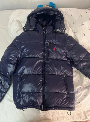 Marinblå glänsig pufferjacka från Polo Ralph Lauren - Säljer en marinblå pufferjacka från Polo Ralph Lauren med blank finish och röd broderad logga på bröstet. Jackan har huva, dragkedja framtill och elastiska muddar. Perfekt för kalla dagar och riktigt snygg streetstil. Pris går att diskutera! 💯🫡