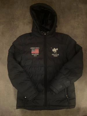 Svart pufferjacka från Vinson Polo Club - Svart pufferjacka med huva från Vinson Polo Club. Jackan har broderade detaljer med amerikansk flagga och texten 'Legend of Sports San Antonio U.S.A' samt Polo Club-logga. Dragkedja framtill och två sidofickor med dragkedja. Perfekt för kalla dagar.