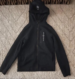 Svart Sail Racing hoodiejacka - Svart hoodiejacka från Sail Racing med broderad logga på bröstet och huvan. Jackan har hel dragkedja framtill, en bröstficka med dragkedja och långärmade ärmar. Mjuk insida i fleece och sportig look, perfekt för chill dagar.