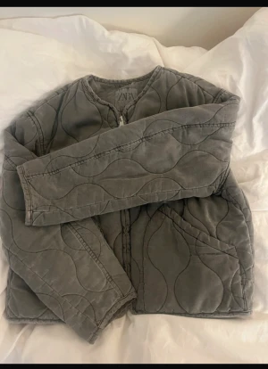 Oliv grön  quiltad jacka från Zara - Quiltad jacka från Zara i oliv grön  med vågigt mönster och rund hals. Jackan har dragkedja framtill och två stora fickor. Materialet är mjukt och känns som bomull, perfekt för dig som gillar chill och enkel stil. 
