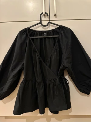 Svart omlottblus med puffärm från Dazy 100 % cotton - Står XL men den i mindre i storlek skulle säga mer som L/M .  Helt i nytt skick/Never worn. 100% cotton / Säljer en svart omlottblus från Dazy med v-ringning och knyt i sidan. Blusen har volangdetaljer vid midjan och långa puffärmar med resår vid ärmslut. Tillverkad i mjuk bomull, perfekt för dig som gillar en chill men ändå snygg look.