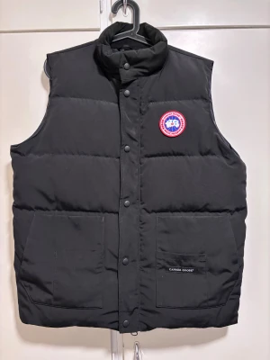 Svart dunväst från Canada Goose XL - Svart dunväst från Canada Goose med hög krage, två stora fickor framtill och klassisk logga på bröstet. Västen har tryckknappar med Canada Goose-gravyr och är perfekt för lager-på-lager. Herrmodell storlek XL, knappt använd! Hel/ren från djurfritt hem!