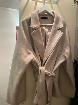 Beige kappa med bälte från Gina Tricot - Snygg beige kappa från Gina Tricot i storlek Xxl . Jackan har breda slag, knytbälte i midjan och långa ärmar. Materialet är mjukt och känns som ullblandning. Perfekt för dig som gillar stilrena och klassiska plagg med modern touch.