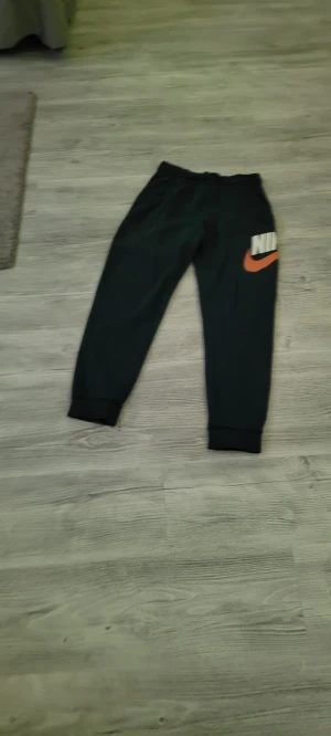 Svarta mjukisbyxor från Nike - Svarta mjukisbyxor från Nike med stor vit logga och orange swoosh på ena benet. Byxorna har resår i midjan och muddar vid bensluten för en skön passform. Perfekta för chill eller träning. Materialet är mjukt och bekvämt.