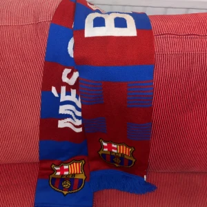 FC Barcelona halsduk röd och blå - Säljer en snygg FC Barcelona halsduk i rött och blått med klubbens emblem broderat i båda ändar. Halsduken har randigt mönster och texten 'Més que un club' i vitt. Perfekt för dig som vill visa ditt stöd på matchen!