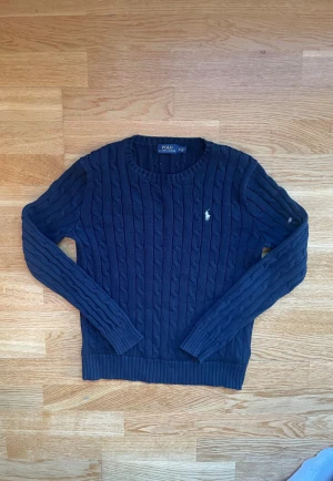 Mörkblå stickad tröja Polo Ralph Lauren - Snygg mörkblå stickad tröja från Polo Ralph Lauren med klassiskt kabelstickat mönster och rund hals. Tröjan har långärmade ribbade muddar och den ikoniska vita logon broderad på bröstet. Perfekt för dig som gillar stilren och tidlös design.