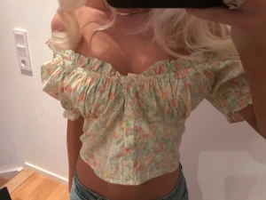 Blommig offshoulder croppad blus - Säljer en jätte söt blommig offshoulder blus i ljusa pastellfärger med volangdetaljer och knappar framtill. Toppen är croppad och har korta puffärmar, perfekt för sommarens alla häng. Materialet känns lätt och luftigt, och mönstret är romantiskt med små blommor i rosa, grön och beige.