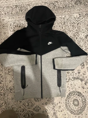 Nike Tech Fleece svart/grå hoodie - Nike Tech Fleece hoodie i svart och ljusgrått med hel dragkedja och huva. Jackan har två fickor med svarta dragkedjor och snyggt kontrasterande design. Perfekt för chill eller träning, med mjukt och flexibelt material.