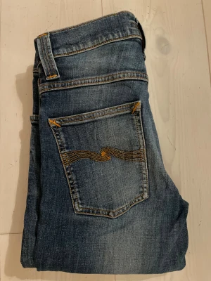 Blå Nudie Jeans  - Snygga blå jeans från Nudie Jeans med slim fit stil och klassisk femficksdesign. Jeansen har gul kontrastsöm och Nudie-logga på bakfickan. Materialet är slitstarkt denim i bomull och passformen är tight hela vägen ner. Perfekt för dig som gillar en schysst look!