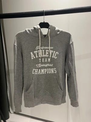 Grå hoodie från PXS Active Sports - Grå hoodie med vit text på bröstet. Tröjan har huva med vita snören och ribbade muddar. Materialet är mjuk bomullsmix och passformen är normal.