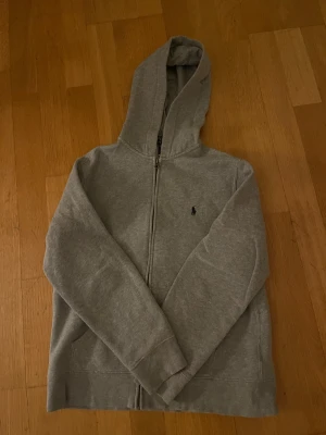 Grå hoodie från Polo Ralph Lauren - Säljer en grå hoodie från Polo Ralph Lauren med dragkedja och huva. Tröjan har långa ärmar, två fickor fram och den klassiska Polo-loggan broderad på bröstet. Mjuk insida och normal passform, perfekt för chill dagar.