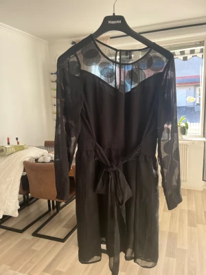 Svart kortklänning från Vero Moda XL - Svart kortklänning från Vero Moda i transparent material med stora prickar på ärmar och upptill. Klänningen har långärmade ärmar, knytband i midjan och rund halsringning. Snyggt flowy tyg som ger en cool look.