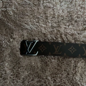 bälte - Snyggt bälte i brunt skinn med klassiskt monogrammönster och silverfärgat spänne. Bältet har beige insida och tydliga logodetaljer. Perfekt accessoar för att lyfta din outfit med en lyxig touch.