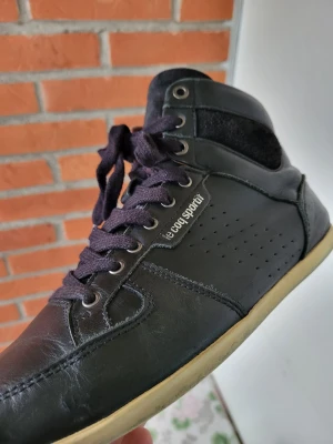 Svarta Le Coq Sportif sneakers high 42 - Svarta high-top sneakers från Le Coq Sportif med snörning och rund tå. Skorna har en ovandel i skinn med detaljer i mocka och perforeringar på sidorna. Yttersulan är beige och insidan är brun. Perfekt för dig som gillar klassisk streetstil.